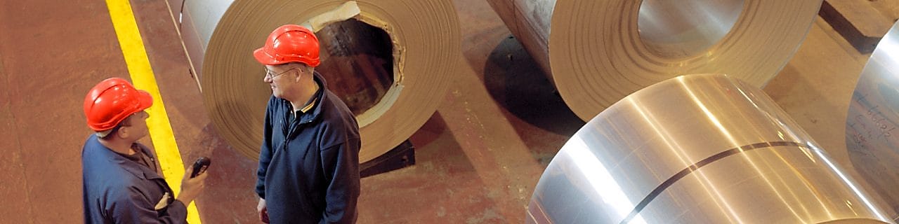 Two factory workers standing next to rolls of steel